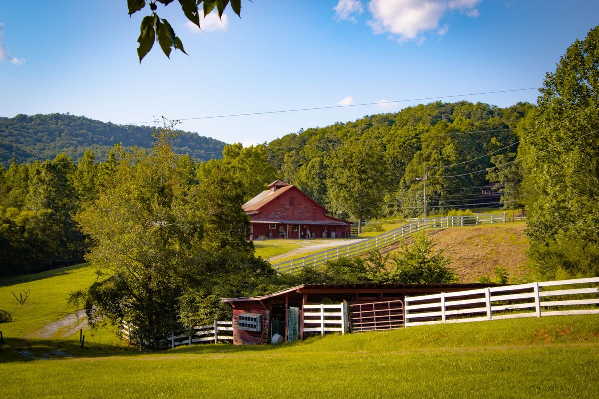 Red barn and white board fences on a green farm below wooded hills
