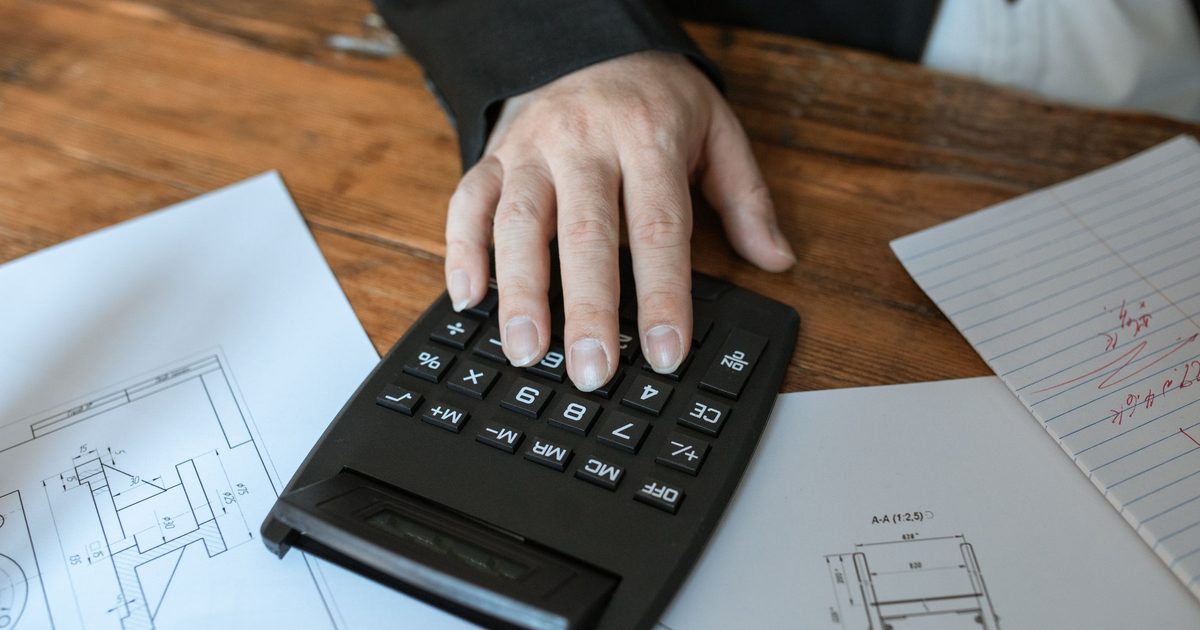 Hand on a calculator beside technical drawings on a wooden desk